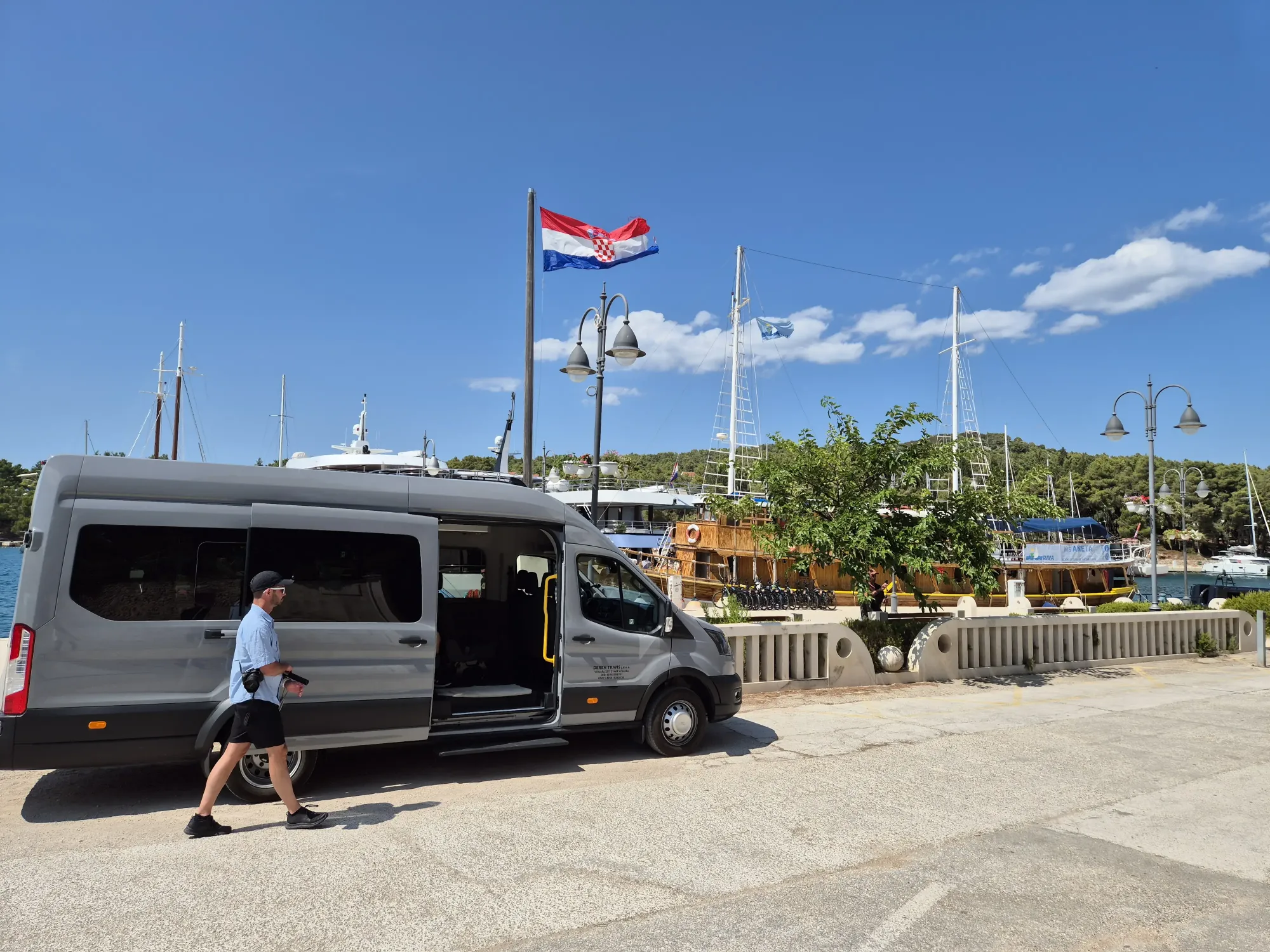 Driver boarding a group transfer minibus on a Hvar harbour promenade with the Croatian flag and yachts