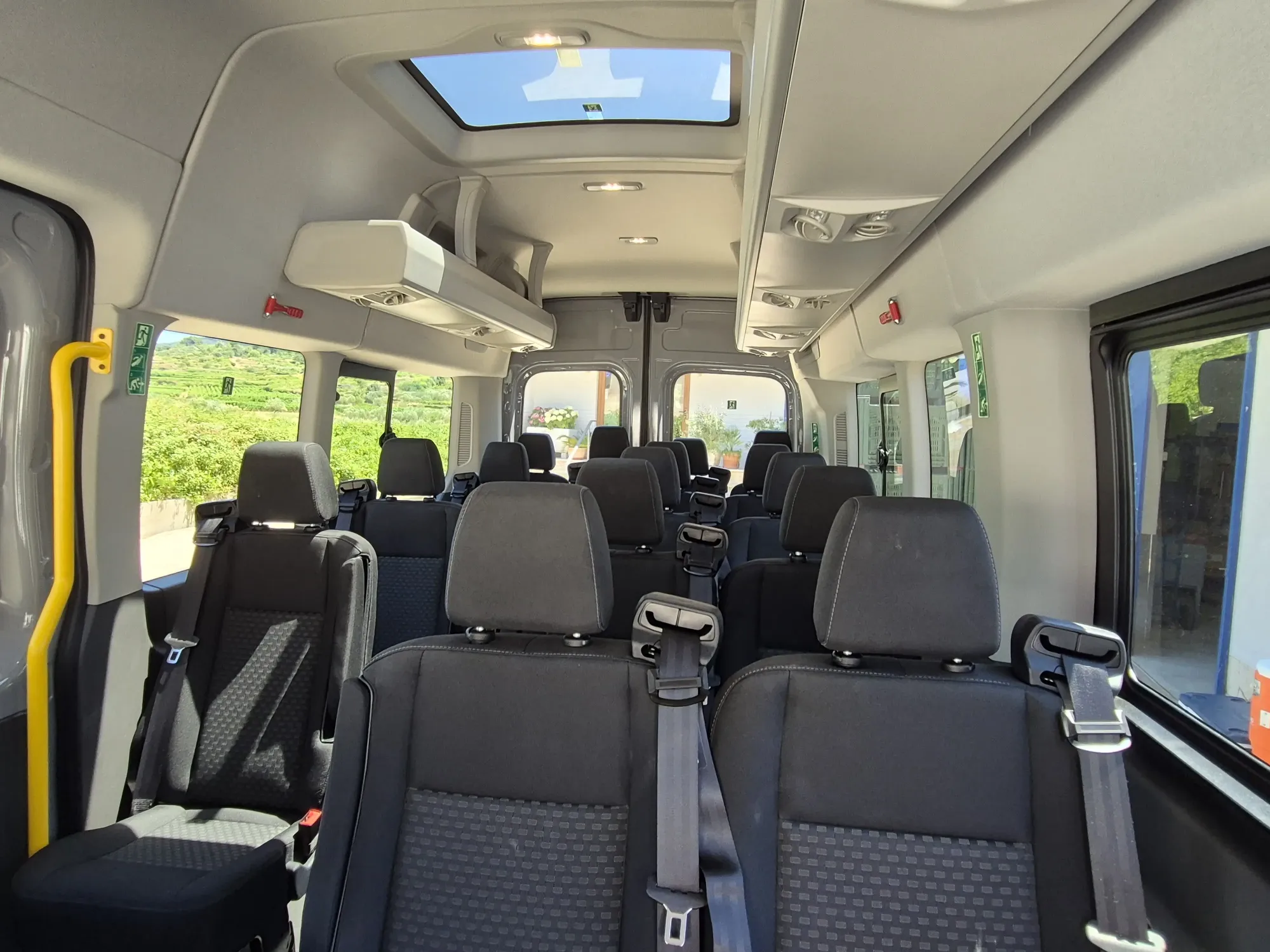 Interior of a group transfer minibus showing rows of seats, seatbelts and a panoramic sunroof
