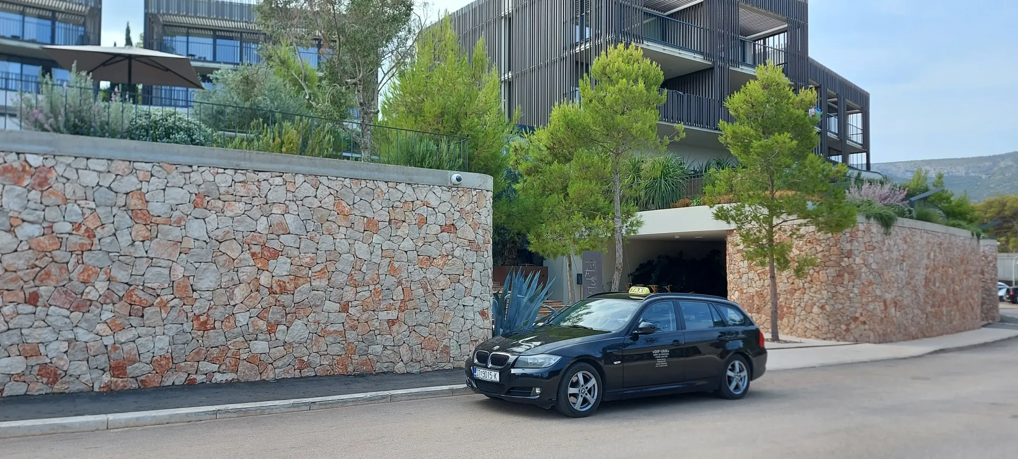 Black standard taxi car parked by a stone wall outside a modern stone hotel on Hvar