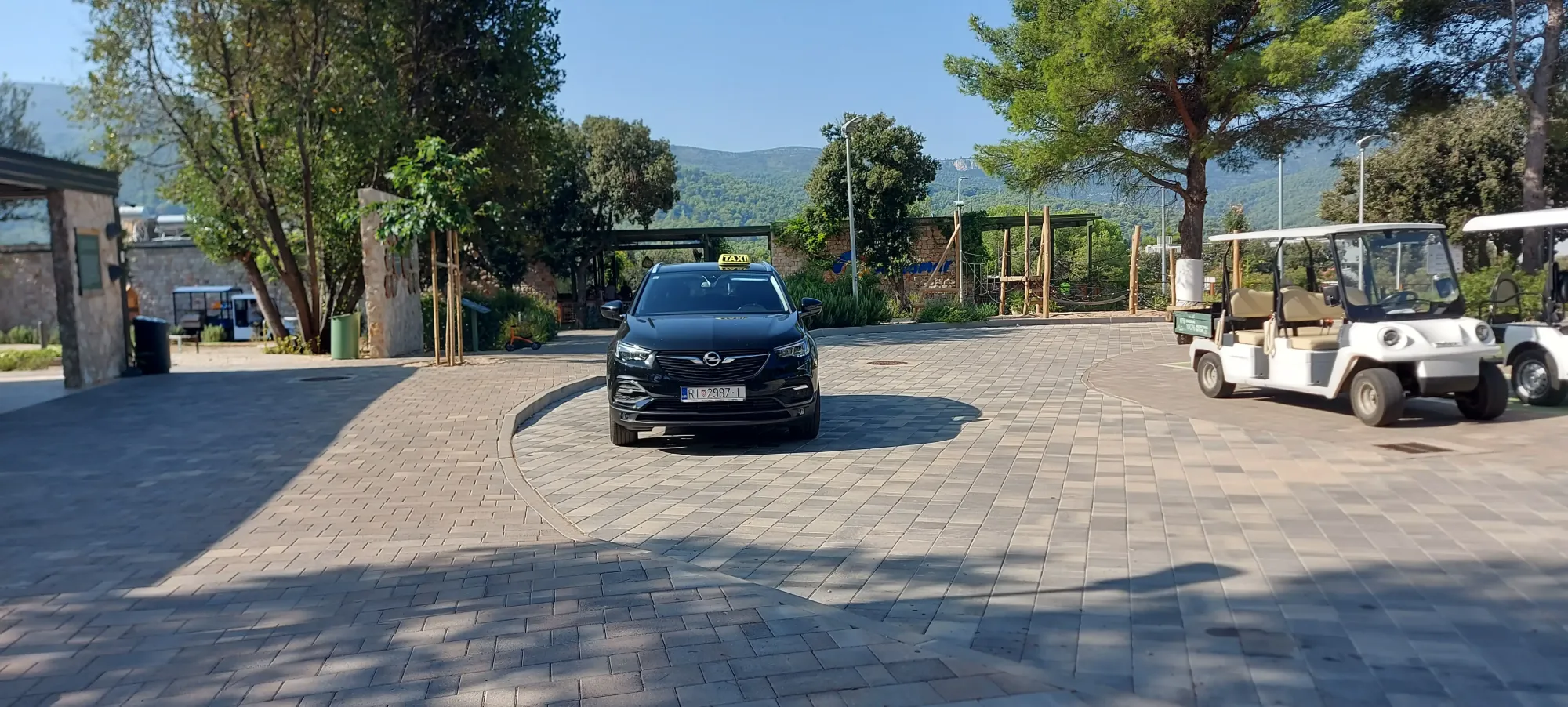 Black estate taxi parked on a paved resort forecourt next to white golf buggies