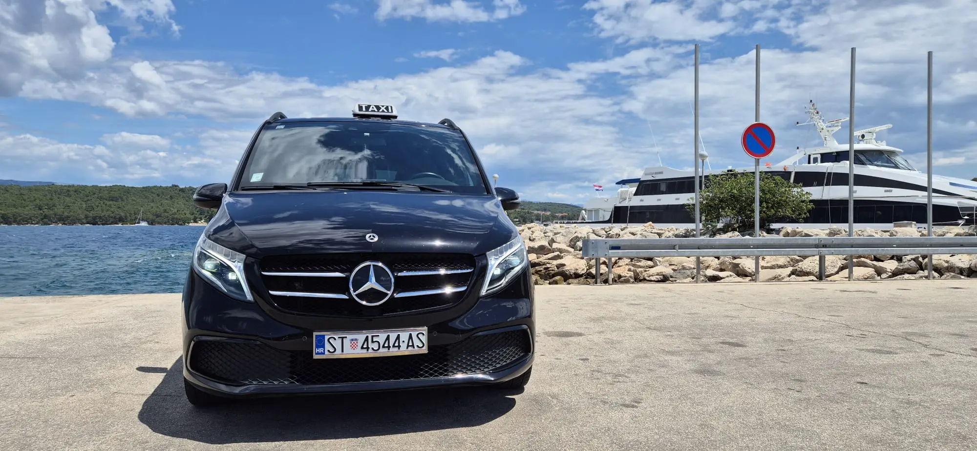 Black luxury van taxi parked front-on at a Hvar harbour with a yacht and the sea behind